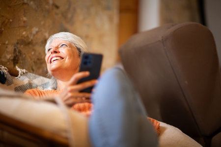 Smiling elderly woman in an orange sweater reclining on a couch, holding a smartphone and enjoying a quiet, relaxed moment at homeの写真素材