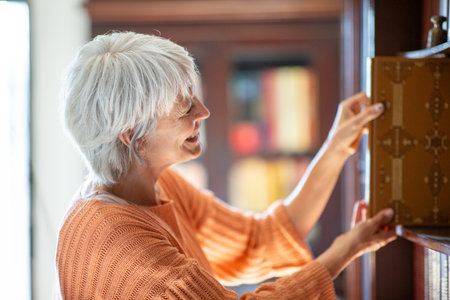 Side profile of a mature woman with short gray hair, smiling and wearing an orange sweater, pulling a book from a library shelfの写真素材