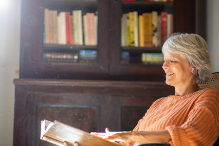 Mature woman with short gray hair, wearing an orange sweater, sitting by a bookshelf and reading a book with a relaxed smileの写真素材