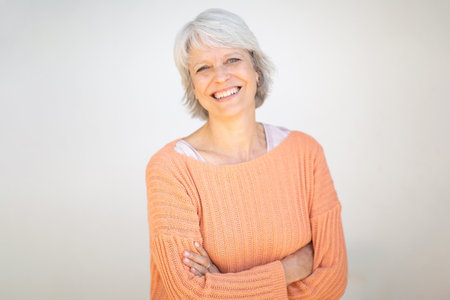 Portrait of a mature woman with short gray hair, wearing an orange sweater and smiling with arms crossed, standing in front of a white backgroundの写真素材