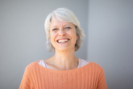 Close-up portrait of a mature woman with short gray hair, smiling warmly and wearing an orange sweater, standing against a light gray backgroundの写真素材