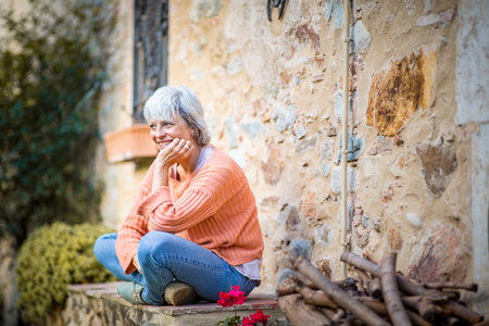 A senior woman with grey hair, wearing an orange sweater and jeans, sits outdoors on a stone step with her chin resting on her handsの写真素材