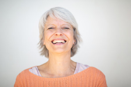 Close-up portrait of a mature woman with short gray hair, wearing an orange sweater, smiling openly in front of a white backgroundの写真素材