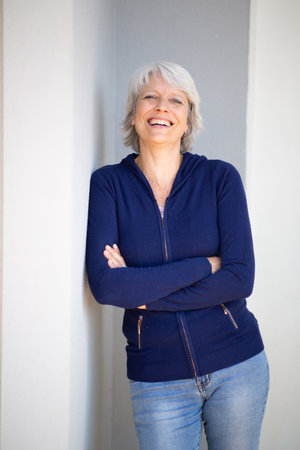 Portrait of a mature woman with gray hair, dressed in a navy blue sweater and jeans, smiling and standing with arms crossed while leaning against a light wallの写真素材