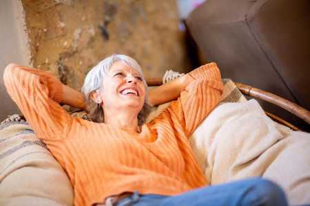 A senior woman with grey hair is seen reclining on a comfortable couch, smiling and looking upの写真素材