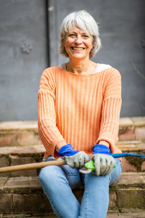 Smiling elderly woman in an orange sweater and jeans sitting on steps outdoors, holding a garden rake and pruning shearsの写真素材