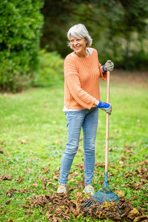 A senior woman with short gray hair, dressed in an orange sweater, jeans, and gloves, is raking leaves in an autumn gardenの写真素材