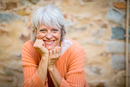 A senior woman with grey hair, dressed in an orange sweater, smiles warmly while resting her chin on...