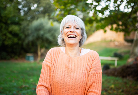 A senior woman wearing an orange sweater smiling warmly while standing in a gardenの写真素材