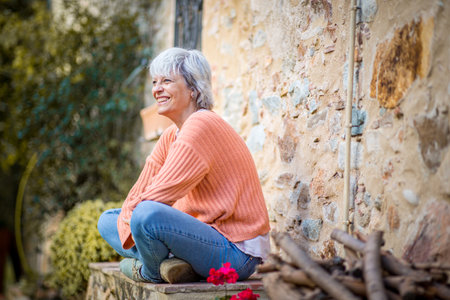 A senior woman with grey hair, dressed in an orange sweater and jeans, sits outdoors on a stone stepの写真素材