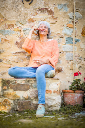 A senior woman with grey hair is sitting outdoors, smiling as she talks on the phoneの写真素材