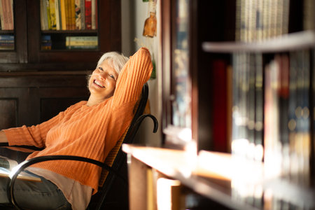 Senior woman in an orange sweater reclining in a chair, smiling and enjoying natural sunlight in a cozy home libraryの写真素材