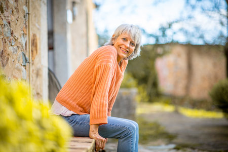 A senior woman with grey hair, wearing an orange sweater and jeans, sits outdoors on a bench, smiling brightlyの写真素材