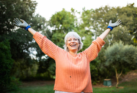 A cheerful elderly woman wearing an orange sweater and gloves, raising her arms with a big smile in a gardenの写真素材