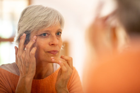 Senior woman in an orange sweater gently applying eye cream while looking at herself in the mirrorの写真素材