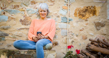 A senior woman with grey hair, wearing an orange sweater and jeans, is sitting outside against a rustic stone wall, holding a smartphoneの写真素材