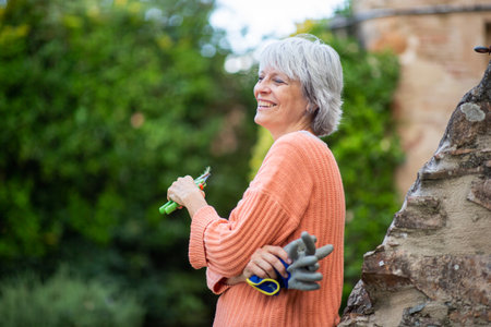 A senior woman with grey hair, wearing an orange sweater, smiles while holding gardening gloves and pruning shears in her backyardの写真素材