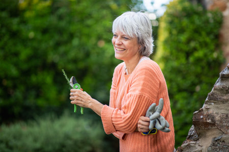 A senior woman with grey hair, wearing an orange sweater, smiles while holding pruning shears and gardening gloves in a peaceful outdoor settingの写真素材