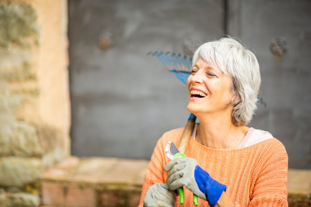A senior woman with short gray hair, wearing an orange sweater and gardening gloves, is laughing while holding pruning shears and a rakeの写真素材