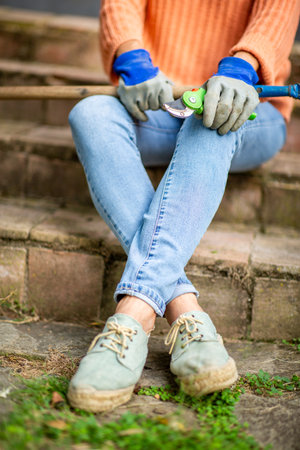 Close-up of a person sitting on stone steps, wearing jeans, green shoes, gardening gloves, and holding pruning shearsの写真素材