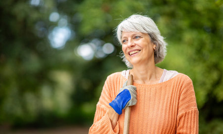 Senior woman with gray hair, wearing an orange sweater and gardening gloves, smiles while holding a rake in an outdoor gardenの写真素材