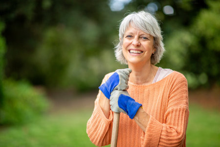 A senior woman with gray hair, wearing an orange sweater and blue gloves, smiling while holding a...