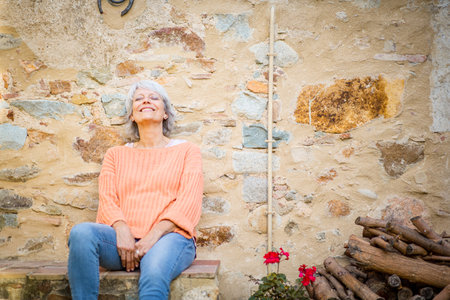 A senior woman with grey hair, wearing an orange sweater and jeans, is sitting outdoors against a rustic stone wallの写真素材