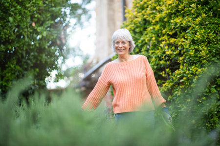 A senior woman with grey hair, wearing an orange sweater, is standing in her garden surrounded by lush greenery and plantsの写真素材
