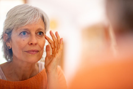 Senior woman in an orange sweater delicately applying skincare around her eyes while gazing in the mirrorの写真素材