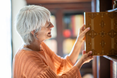 Side profile of a mature woman with short gray hair, smiling and wearing an orange sweater, as she pulls a large book from a library shelfの写真素材