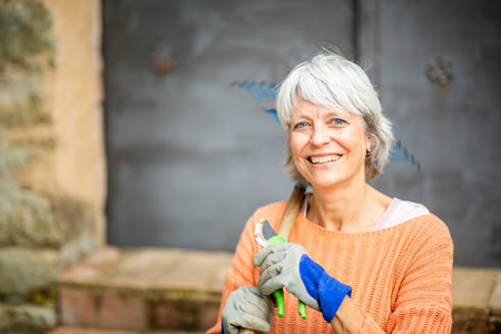 Senior woman with short gray hair, wearing an orange sweater and gardening gloves, smiling while holding pruning shears and a rakeの写真素材