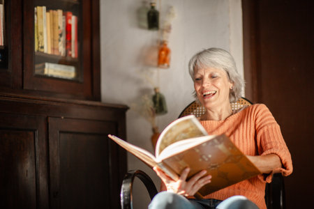 Mature woman with short gray hair, wearing an orange sweater, laughing while reading a book in a cozy home environmentの写真素材