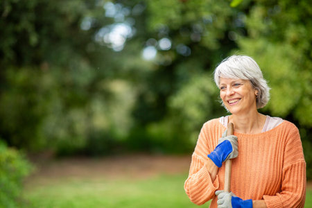A senior woman with short gray hair, wearing an orange sweater and gloves, is smiling while holding a rake in a gardenの写真素材