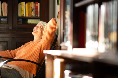 Senior woman in an orange sweater leaning back in a chair and smiling, enjoying the natural sunlight in a home library settingの写真素材