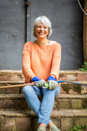 Smiling elderly woman in an orange sweater and jeans sitting on stone steps outdoors, holding a garden rake and pruning shearsの写真素材