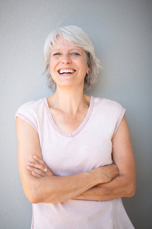 Close-up portrait of a mature woman with short gray hair wearing a light pink shirt, standing with arms crossed and smiling directly at the camera against a gray backgroundの写真素材