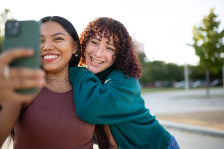Two women standing outdoors, smiling and hugging while taking a selfie with a smartphone. One woman holds the phone as they pose together in a sunny park with trees and an urban backgroundの写真素材