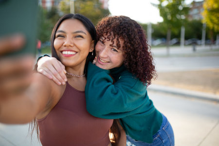 Two women smiling and posing for a selfie together outdoors in a park. One woman holds the smartphone while the other hugs her from behind with trees and an urban backdropの写真素材