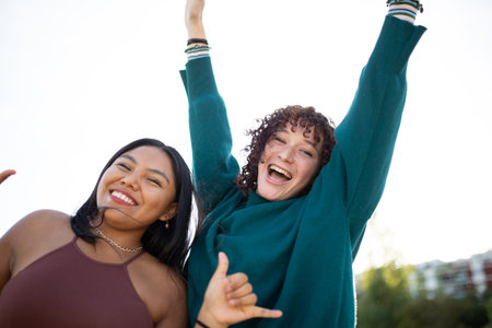 Two joyful young women celebrating with raised arms while posing outdoors on a bright dayの写真素材