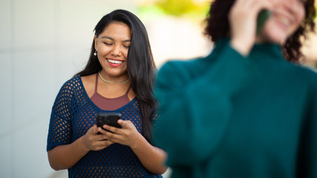 A Latin woman smiles while looking at her phone outdoors. Another woman is in the foreground, laughing and holding a phone to her earの写真素材