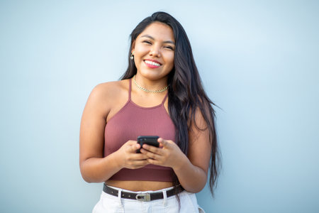 A smiling woman with long dark hair, wearing a gold chain necklace and a brown halter top, holding a smartphone and looking directly at the cameraの写真素材