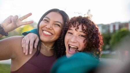 Two women laughing and taking a selfie outdoors, with one flashing a peace sign. They are hugging and enjoying a fun moment in a sunny parkの写真素材
