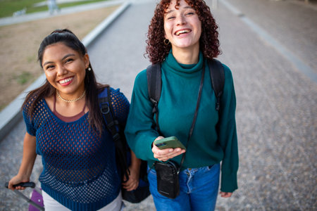 Two happy young women walking outdoors, one holding a smartphone and the other pulling a purple suitcase. They are smiling and enjoying their time together in an urban park settingの写真素材