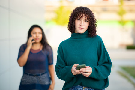 Two young women walking outdoors, one focused on her phone in the foreground while the other is talking on her phone in the backgroundの写真素材