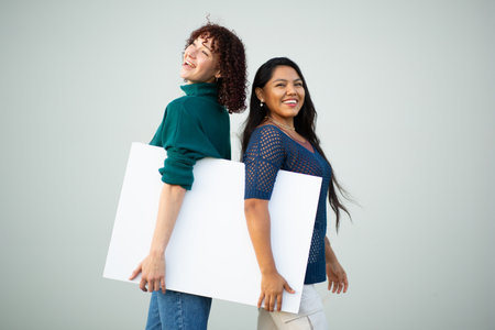 Two smiling women standing back-to-back and holding a blank white sign together with a neutral gray backgroundの写真素材