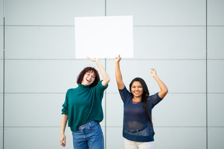Two women standing and celebrating while holding a blank white sign above their headsの写真素材