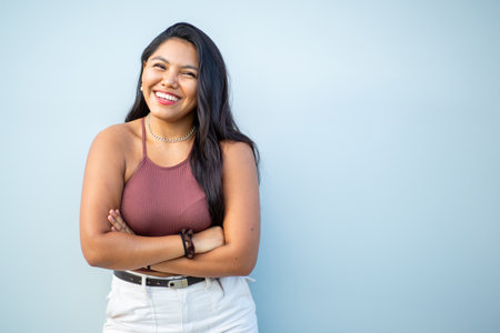 Woman in a sleeveless top and white pants, smiling and standing with her arms crossed, in front of a light blue backgroundの写真素材