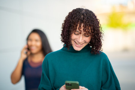 Close-up of a smiling young woman using her smartphone while walking outdoors, with another woman talking on the phone in the backgroundの写真素材