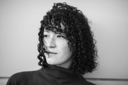 A black and white close-up portrait of a woman with curly hair and freckles, wearing a turtleneck sweater and looking to the sideの写真素材