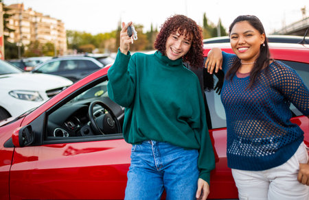 Two women standing beside a red car, smiling happily. One of them is holding car keys, appearing to celebrate a car purchase or new driving experienceの写真素材